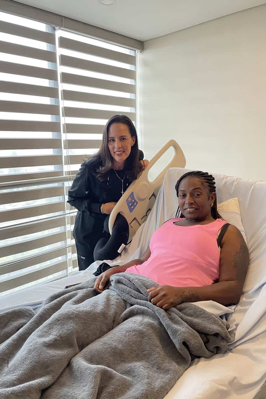 A woman in a hospital chair with another woman standing beside her, both smiling at the camera.