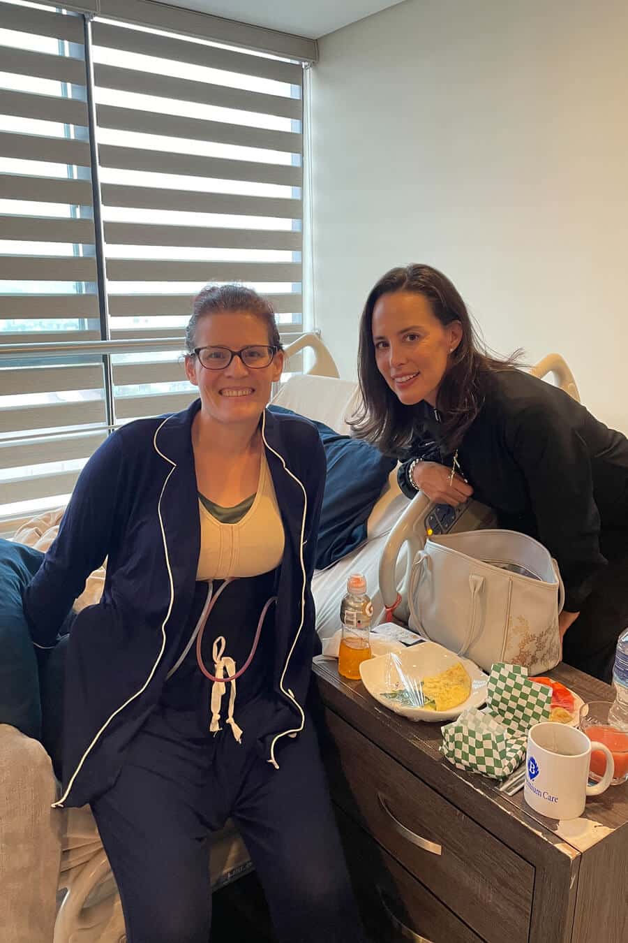 Three women in a hospital setting, one in a wheelchair, all smiling and enjoying a meal together.