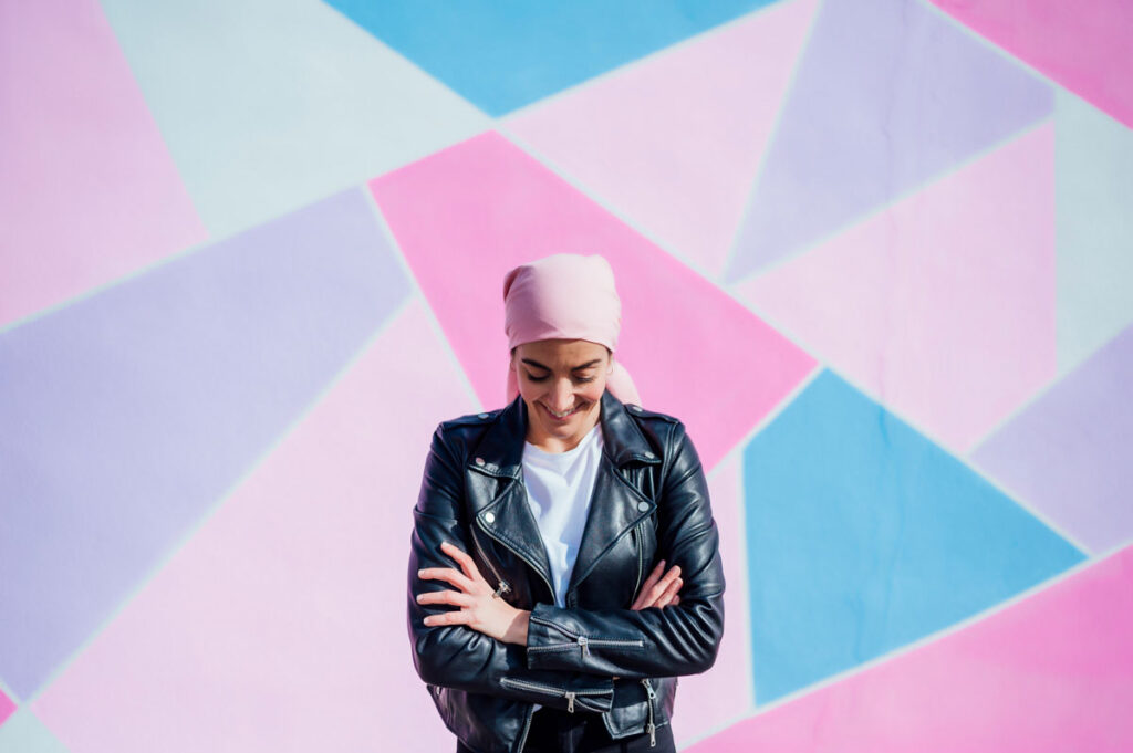 Smiling woman wearing a headscarf, a white shirt, and a black leather jacket, standing in front of a colorful geometric background.