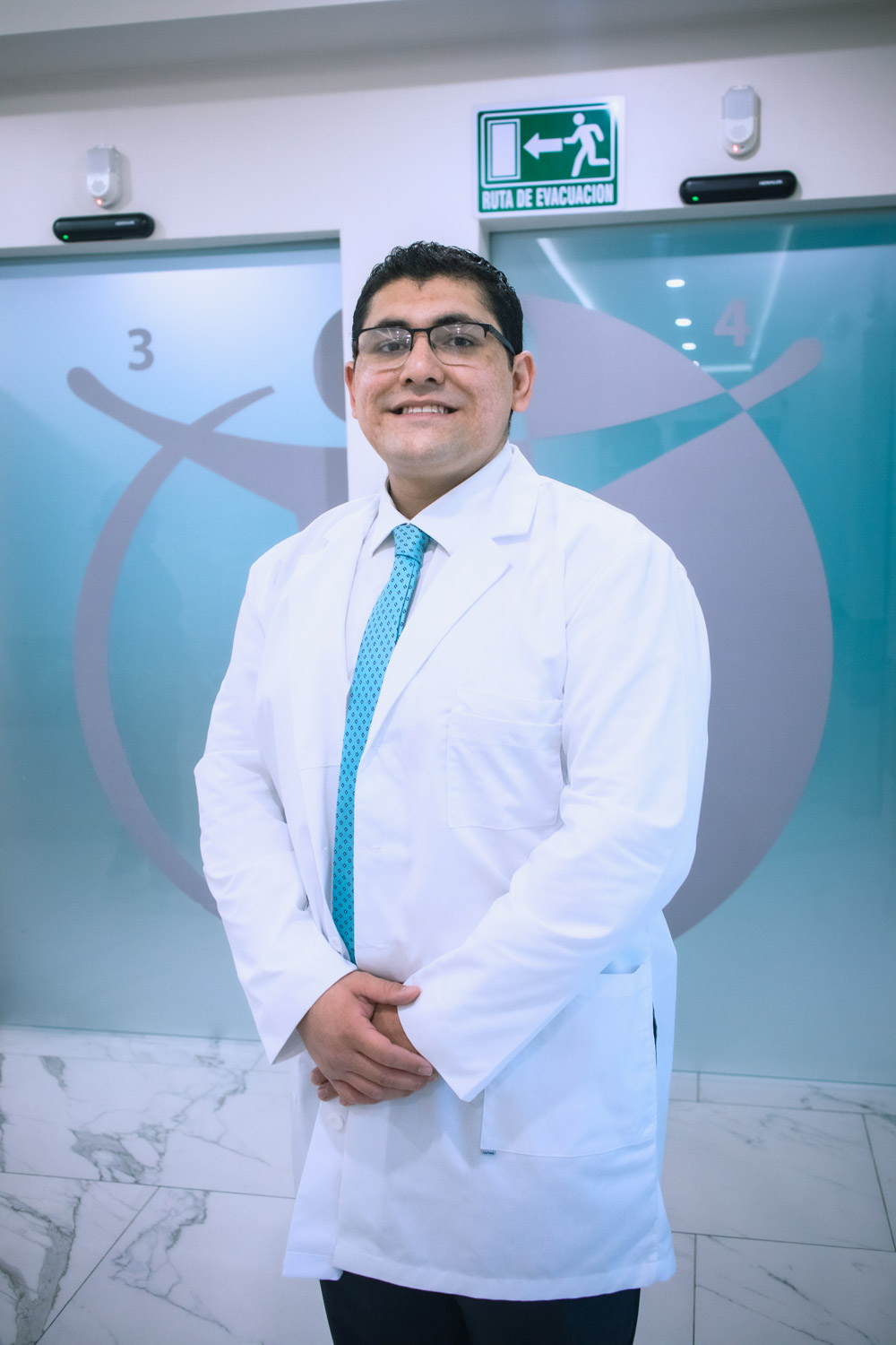 Portrait of a male doctor wearing a white lab coat, smiling, with a sign indicating an evacuation route in the background.