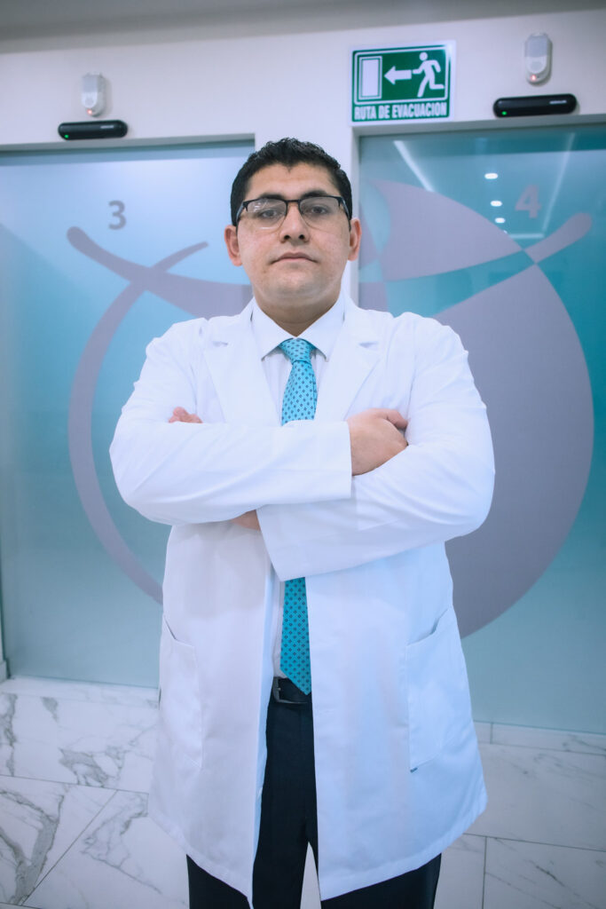 Portrait of a male doctor wearing a white lab coat, standing with arms crossed, with a sign indicating an evacuation route in the background.