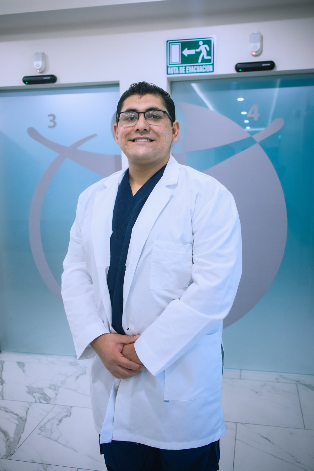 Portrait of a male doctor wearing a white lab coat, smiling, with a sign indicating an evacuation route in the background.