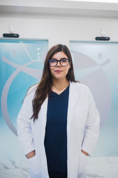 Jlp-tania-01 Doctor in a white lab coat standing and smiling in a medical office, with medical symbols on the frosted glass background.